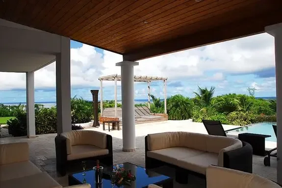 A luxurious covered terrace with dark-wood paneled ceiling and comfortable lounge seating opens to the pool area, pergola with daybeds, lush tropical greenery, and distant ocean views.