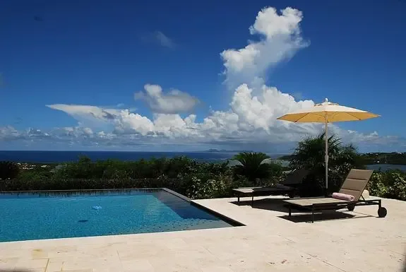 Lounge by the infinity pool on a stone terrace with sun umbrella, overlooking lush tropical landscaping and the sparkling ocean under blue skies.