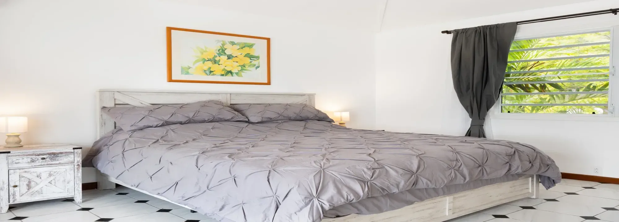Bright bedroom with a king-size bed featuring gray textured bedding, white rustic wooden furniture, and a view of tropical greenery through the window