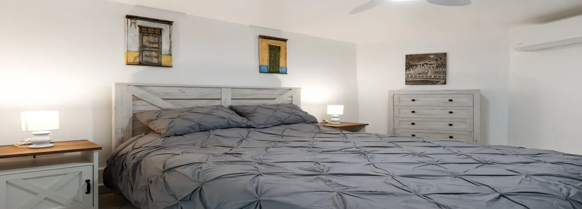 Bright bedroom featuring a large bed with grey linens, white wooden furniture, and decorative wall art