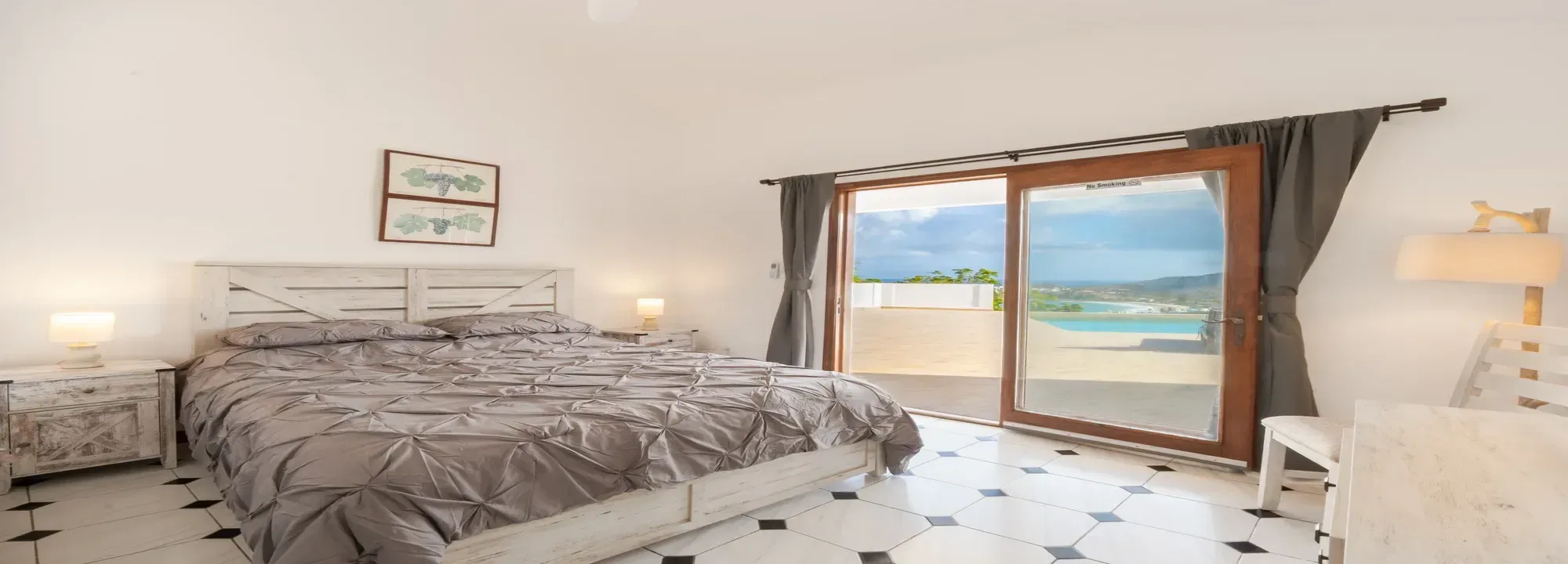 Bright modern bedroom with a grey duvet, white rustic furniture, and sliding glass doors opening to a terrace with turquoise ocean view