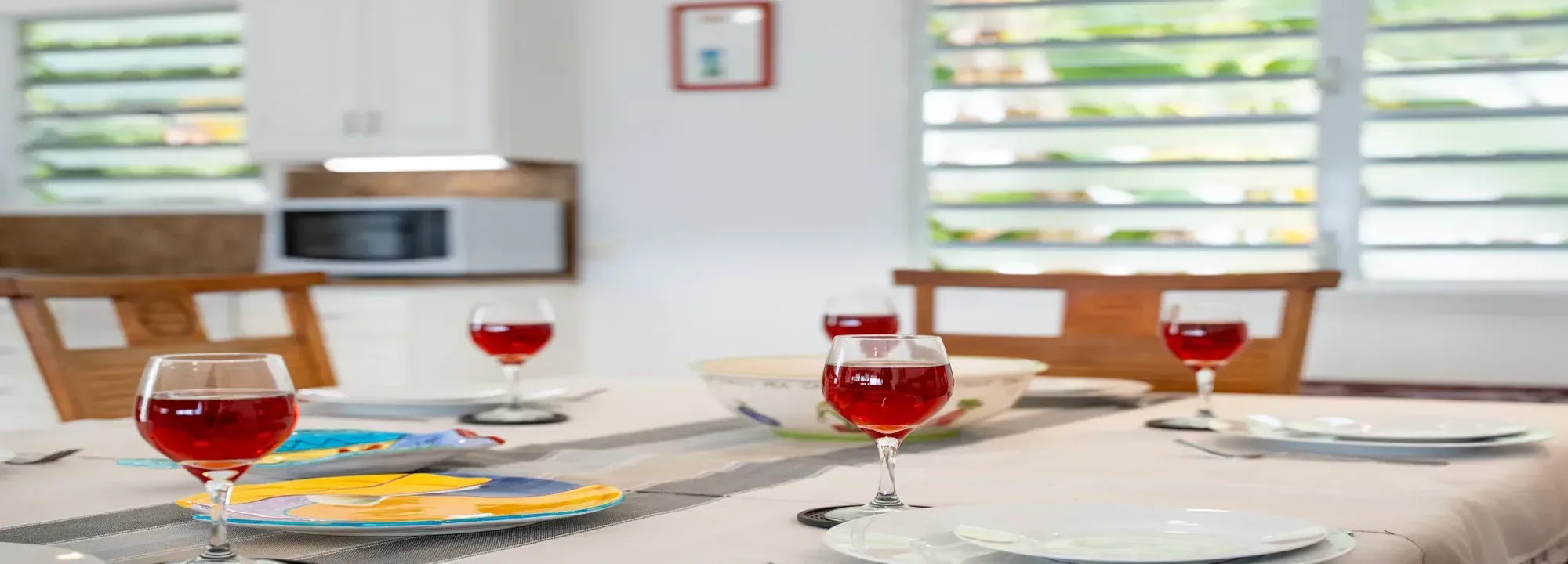 Bright dining room with wine glasses and decorative plates on a table, featuring a modern white kitchen and louver windows in the background