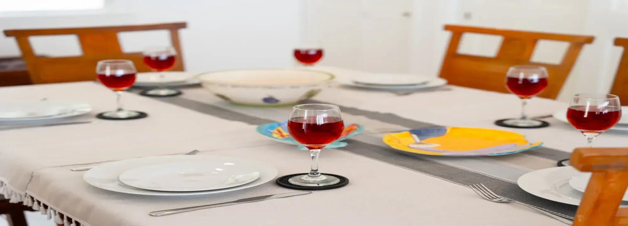 White dining table set with red wine glasses, white ceramic plates, colorful decorative dishes, and wooden chairs in a bright interior