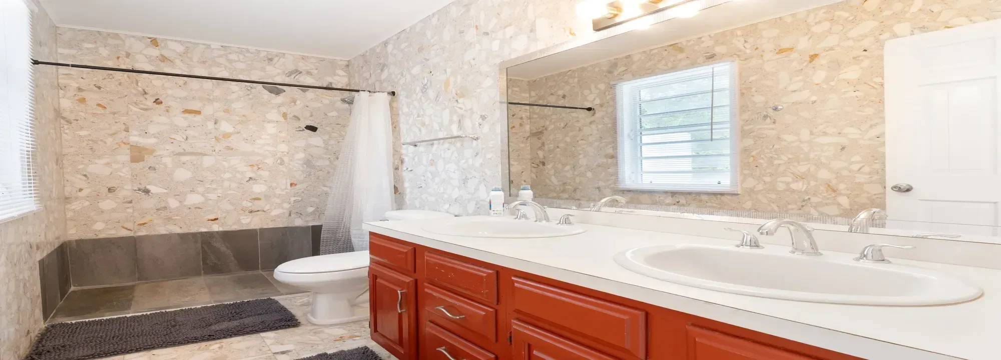Bright bathroom featuring a terracotta double vanity with white sinks, marble-style tiled walls, and a walk-in shower