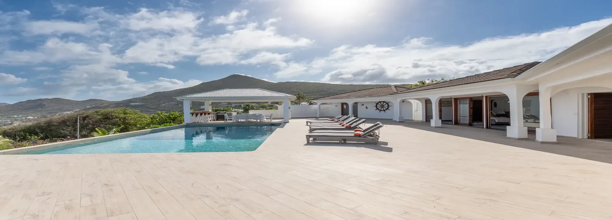 Luxurious modern villa featuring a large beige terrace, infinity swimming pool with lounge chairs, and a white outdoor gazebo overlooking green mountains