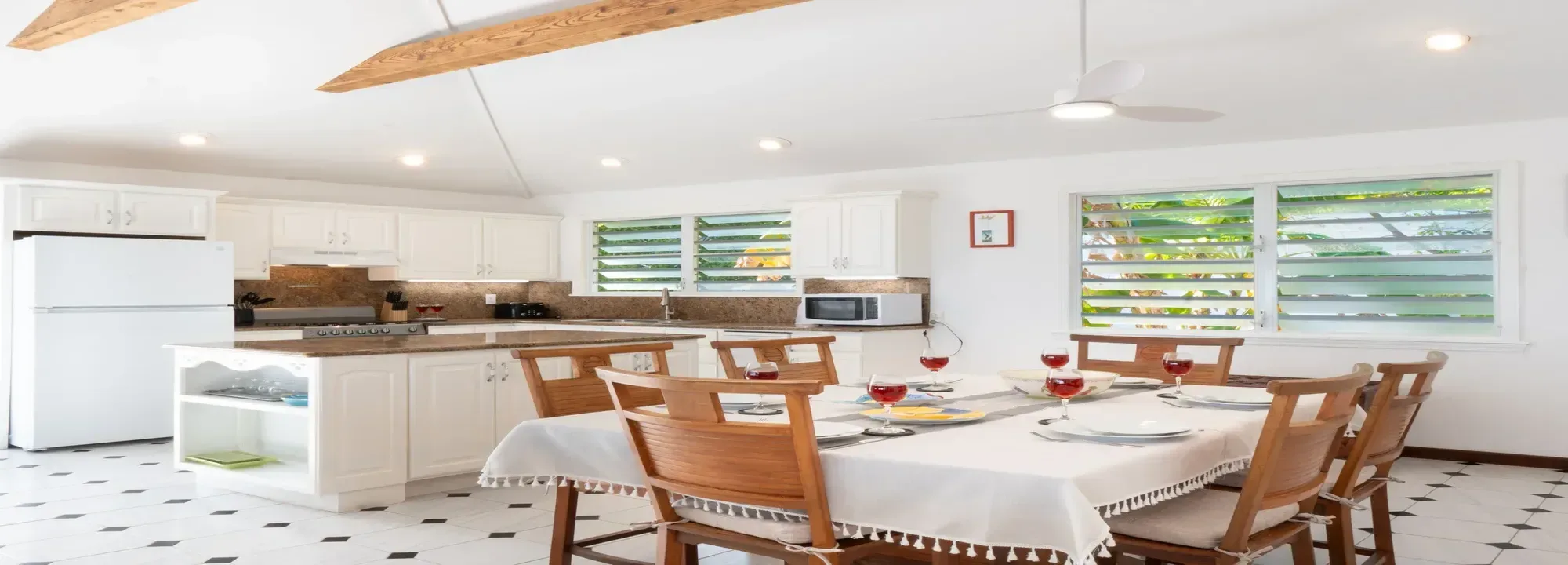 Bright open-concept kitchen and dining room featuring white cabinetry, wooden ceiling beams, and tropical views through louvered windows