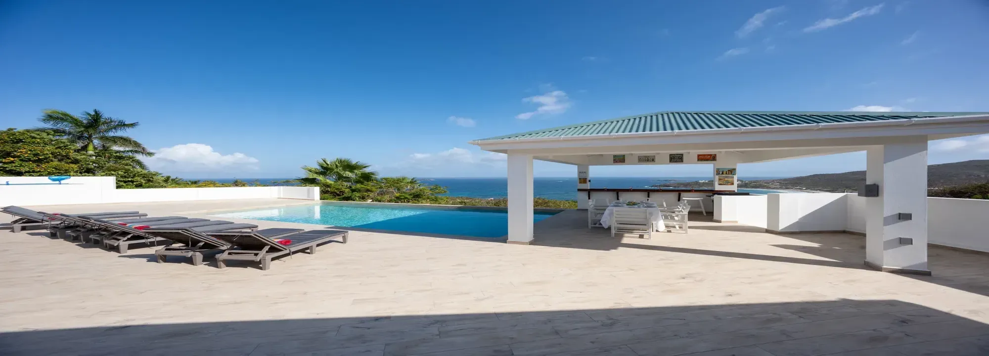 Luxury villa terrace featuring a sparkling blue swimming pool, grey lounge chairs, and a white covered outdoor dining area overlooking the ocean