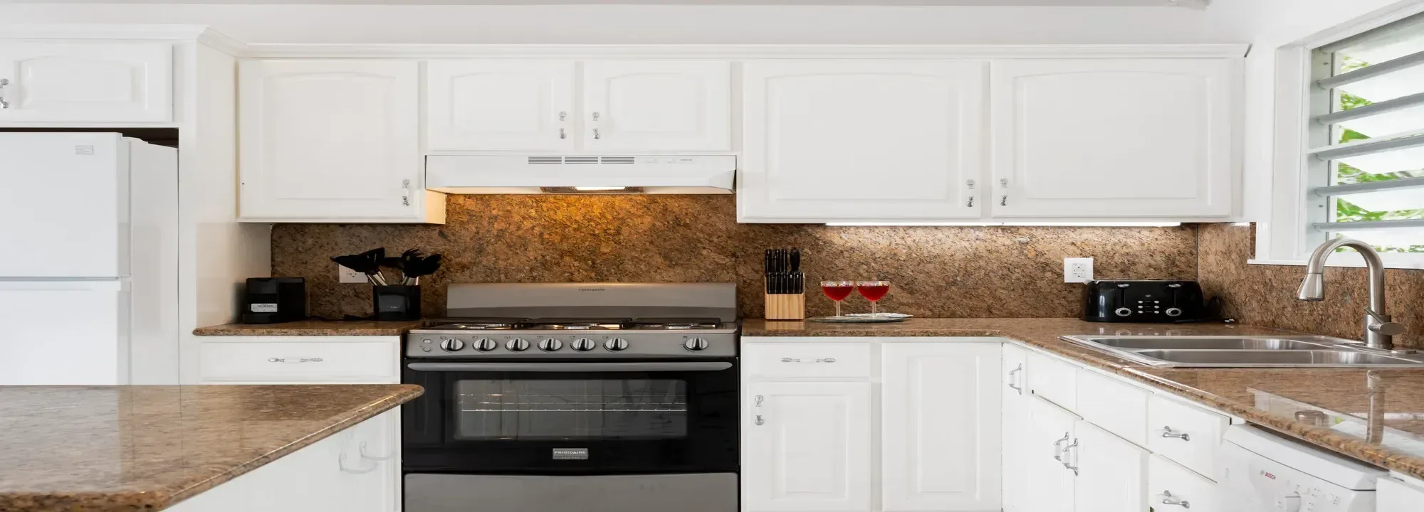 Bright modern kitchen featuring white cabinets, brown granite countertops, stainless steel appliances, and a window with a view of greenery