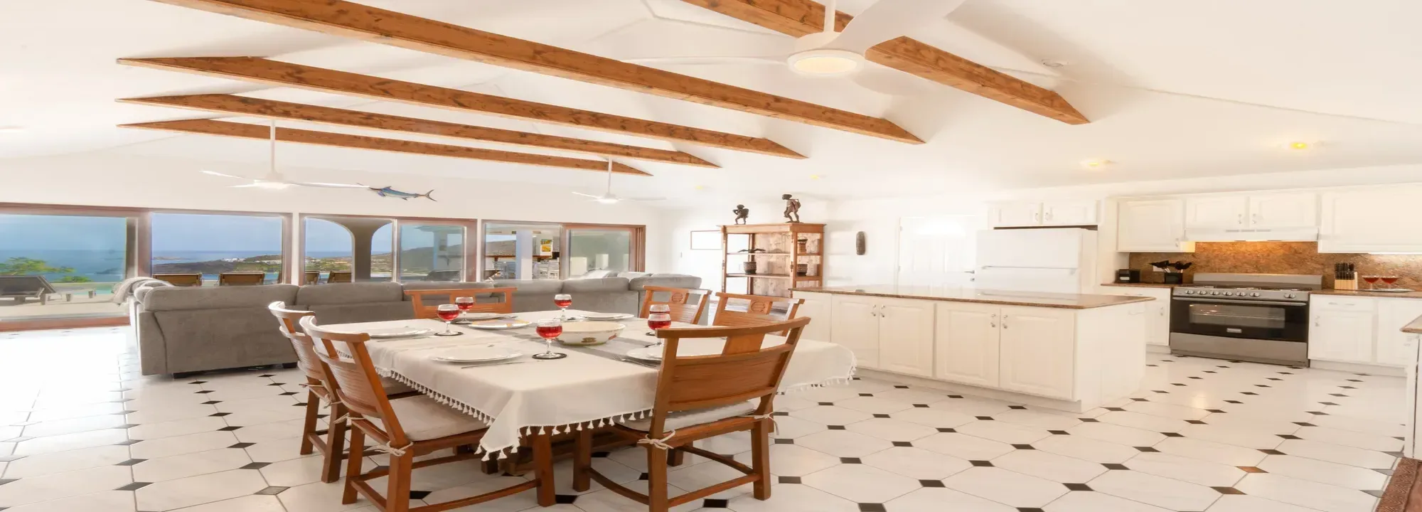 Bright open-concept interior featuring a dining area, gray sectional sofa, white kitchen, and panoramic ocean views