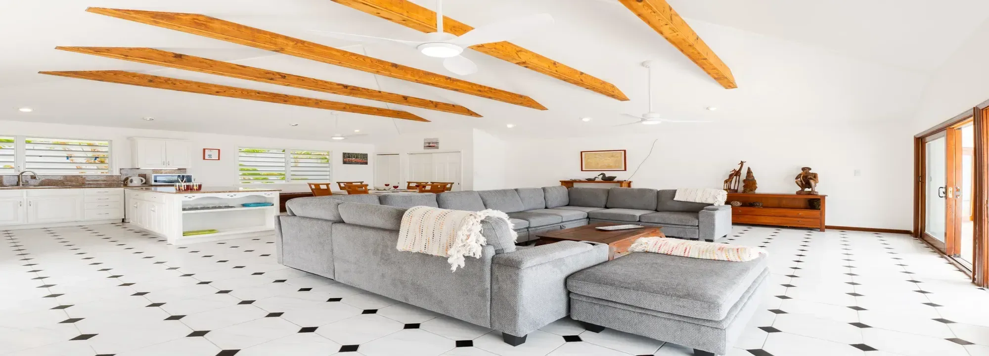 Bright open-concept living room and kitchen featuring grey sectional sofa, white tiled floors, and rustic wooden ceiling beams
