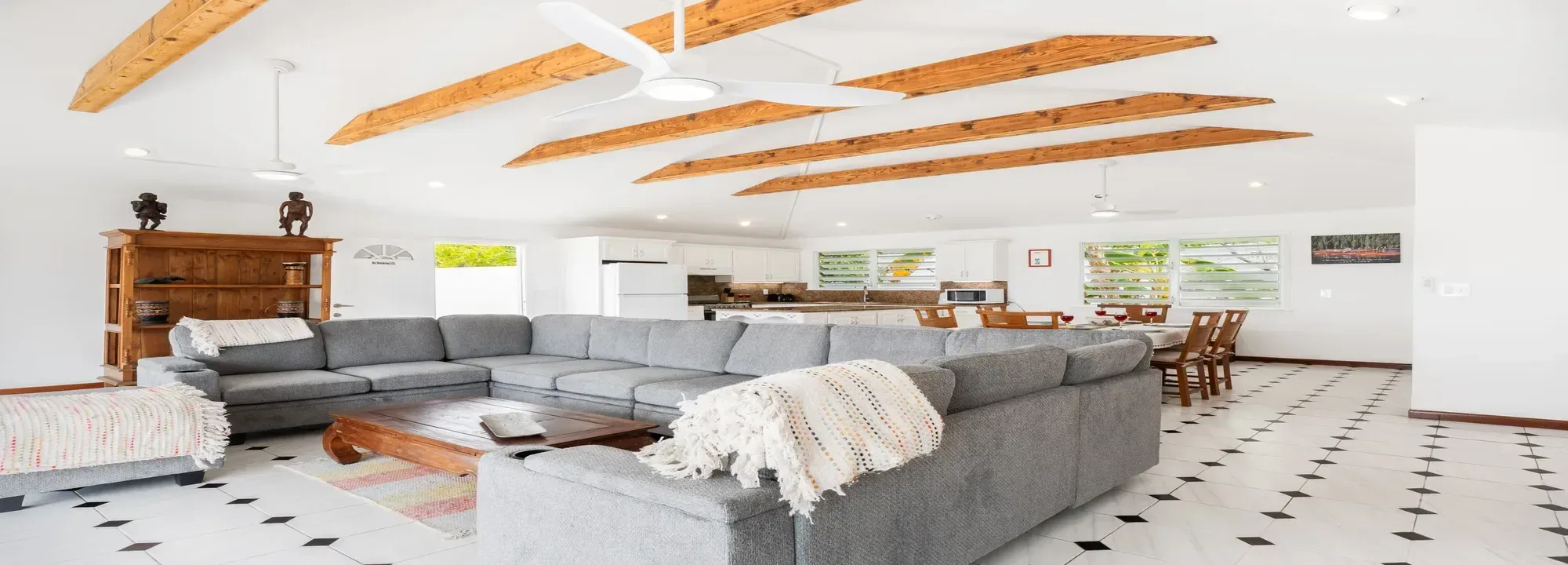 Spacious open-concept living room and kitchen featuring a large grey sectional sofa, rustic wooden beams on white ceiling, and tropical greenery views