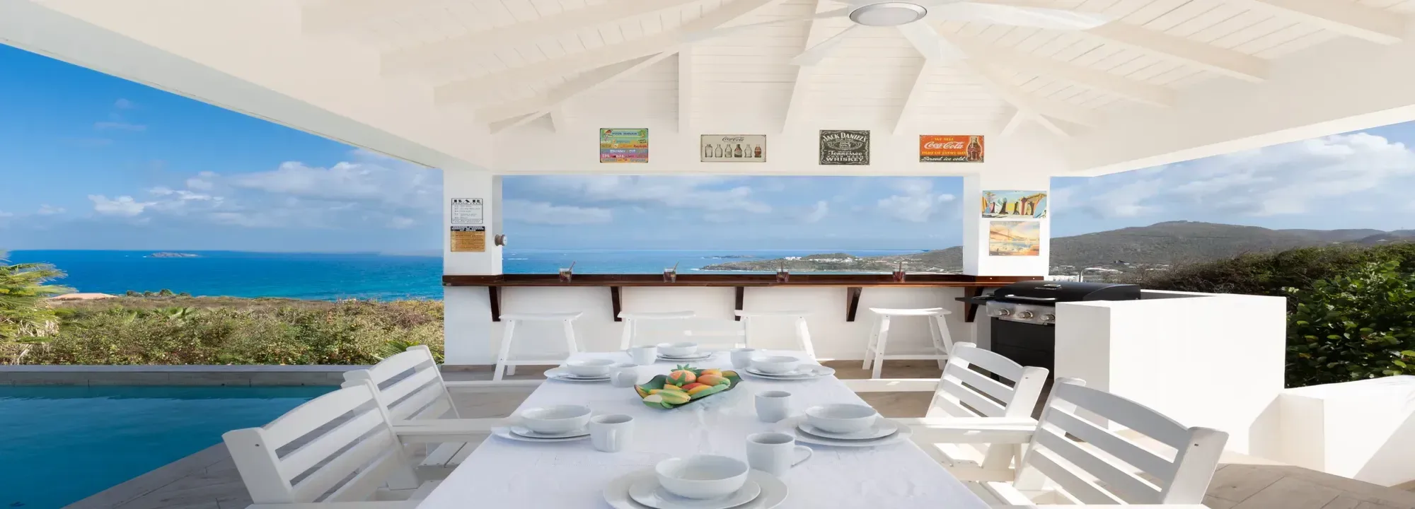 Bright outdoor dining area and bar with white furniture, dining table, and panoramic view of the turquoise ocean and green hills