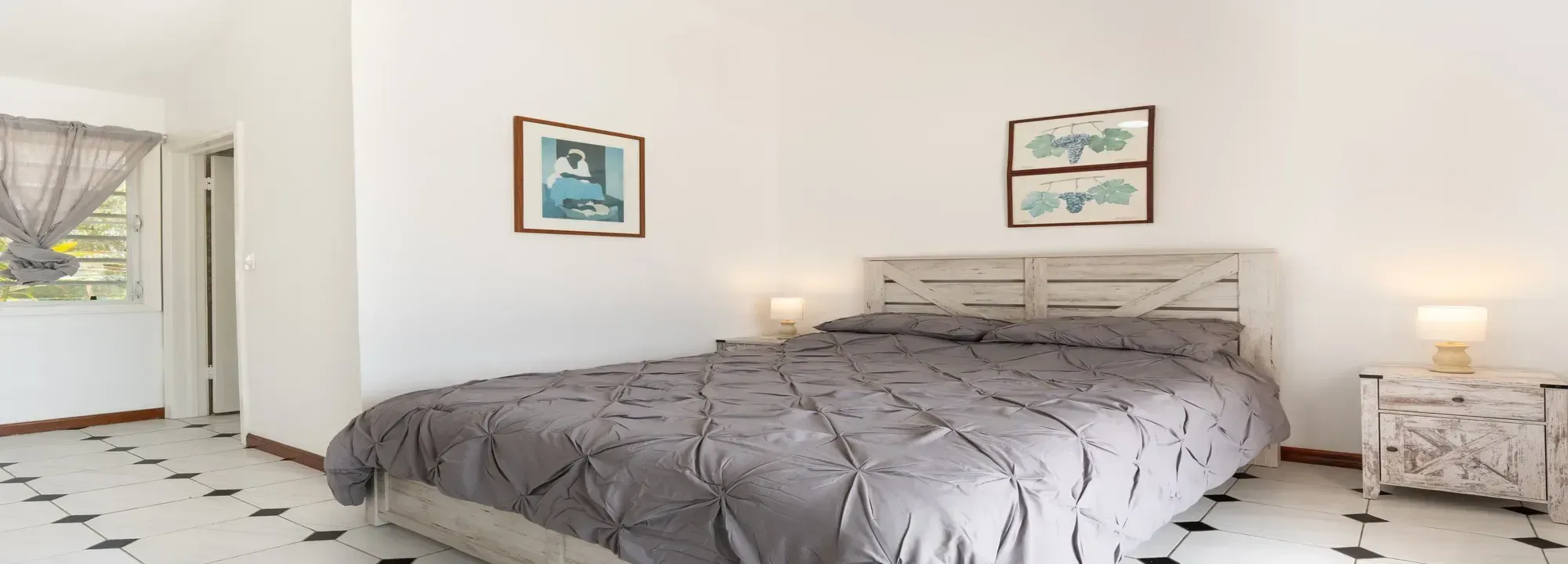 Bright bedroom with a large bed, grey duvet, white rustic wooden furniture, and white tiled floors