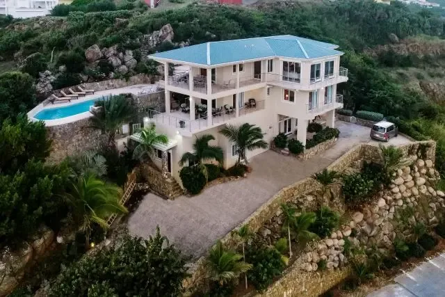 The full scope of Villa Bougainvillea revealed: a three-story Caribbean estate with kidney-shaped pool, lounge chairs, and gravel driveway with dark vehicle. Stone retaining walls and tropical plantings embrace the property, all overlooking the sparkling Caribbean Sea.                                                                                                                                                                                                                                             +
 