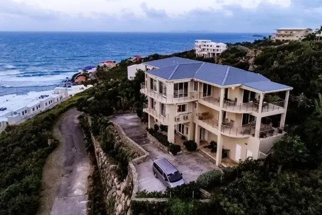 A stunning drone perspective of Villa Bougainvillea's three-story structure, stone retaining walls, and paved driveway. The silver van provides scale while the ocean stretches to the horizon, showcasing this hillside estate's commanding position in Dawn Beach Estates.                                                                                                                                                                                                                                            +
 