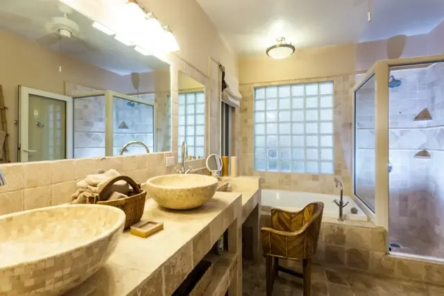 Spa-inspired luxury defines this bathroom, where dual stone vessel sinks anchor a long granite countertop. A glass-enclosed shower with multiple showerheads, freestanding soaking tub, and elegant glass block windows create a private retreat in earth tones worthy of a five-star resort.                                                                                                                                                                                                                             +
 
