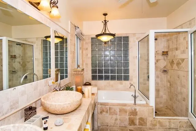 Indulge in spa-level luxury with a large round vessel sink, freestanding soaking tub, and walk-in glass shower. An elegant chandelier illuminates the space while glass block windows provide privacy without sacrificing natural light—a serene Caribbean sanctuary.                                                                                                                                                                                                                                                        +
 