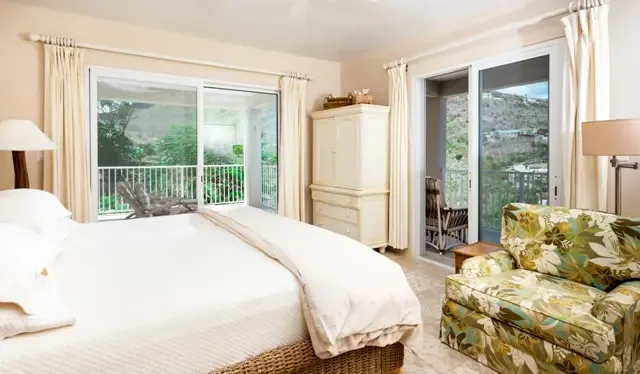A peaceful bedroom retreat featuring a woven bed frame and charming floral armchair, with sliding glass doors opening to views of St. Maarten's lush hillside. The wooden armoire and natural wicker accents complement the island's tropical aesthetic.                                                                                                                                                                                                                                                                 +
 