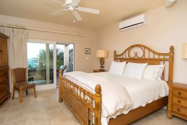 A refined bedroom featuring a carved wooden headboard and crisp white bedding, with two nightstands and a dresser complementing the island aesthetic. Sliding doors and windows reveal palm trees and the Caribbean Sea, bringing St. Maarten's beauty indoors.                                                                                                                                                                                                                                                           +
 