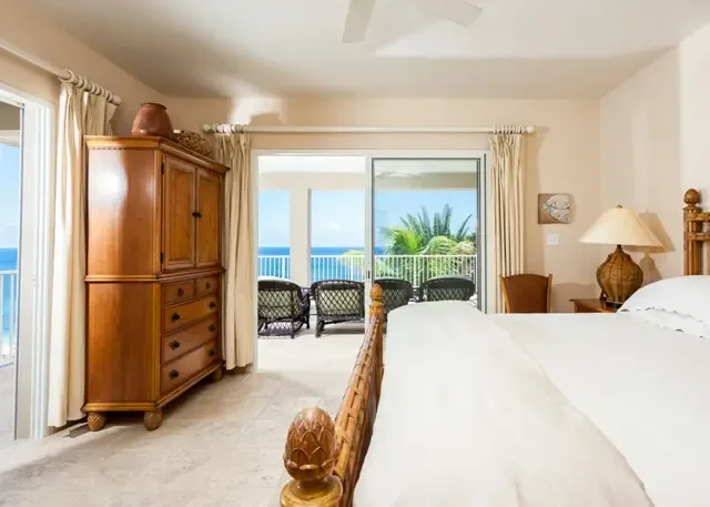 A serene master bedroom featuring a elegant rattan bed frame, wooden armoire, and private balcony access. Sliding glass doors open to reveal the sparkling ocean beyond, while two wicker chairs on the balcony offer the perfect spot to enjoy St. Maarten's coastal beauty at dawn.                                                                                                                                                                                                                                      +
 