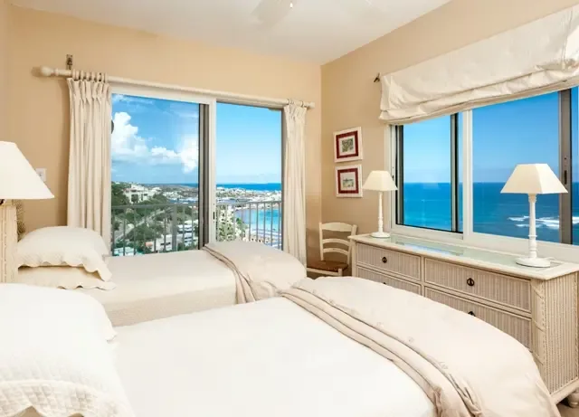 Sunlight floods this inviting bedroom through floor-to-ceiling windows, showcasing breathtaking views of the ocean and coastal town below. Two comfortable beds with crisp white bedding, a dresser with warm lamps, and curated wall art create a relaxing Caribbean escape.                                                                                                                                                                                                                                                +
 