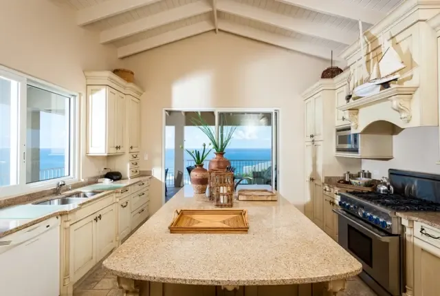 A gourmet kitchen designed for entertaining, featuring a large central island with granite countertop, stainless steel gas cooktop, and all modern appliances. Ocean views through sliding doors inspire culinary creativity, while a model ship and terracotta vase add Caribbean character.                                                                                                                                                                                                                               +
 