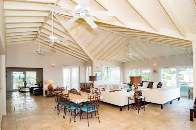 The heart of Villa Bougainvillea: an open living and dining area featuring a white sectional sofa and dining table set for six with blue-cushioned chairs. Multiple ceiling fans and floor lamps provide comfort, while sliding doors frame views of the outdoor terrace and Caribbean landscape.                                                                                                                                                                                                                           +
 