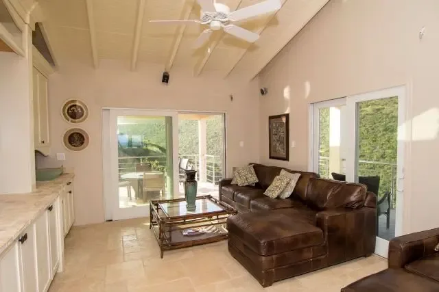 A comfortable living area anchored by a dark brown leather sectional and glass coffee table, with white kitchen cabinets visible beyond. Decorative wall plates and art above the sofa add island character, while sliding doors open to patio views of St. Maarten's lush greenery.                                                                                                                                                                                                                                      +
 