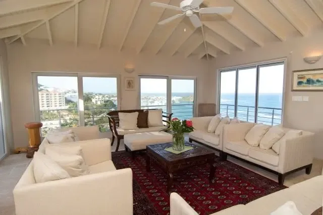 Bright and airy living room with multiple light sofas arranged around a dark wooden coffee table adorned with pink flowers. A red-patterned area rug adds warmth, while large sliding doors frame sweeping ocean views—perfect for indoor-outdoor Caribbean living.                                                                                                                                                                                                                                                          +
 