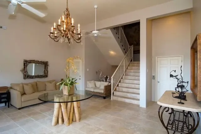 An open-concept living space featuring a beige sofa and wicker armchair around a glass coffee table. A large mirror with rattan frame, elegant chandelier, and ceiling fans accentuate the high ceilings, while a black metal bird sculpture adds artistic Caribbean flair near the staircase.                                                                                                                                                                                                                                +
 