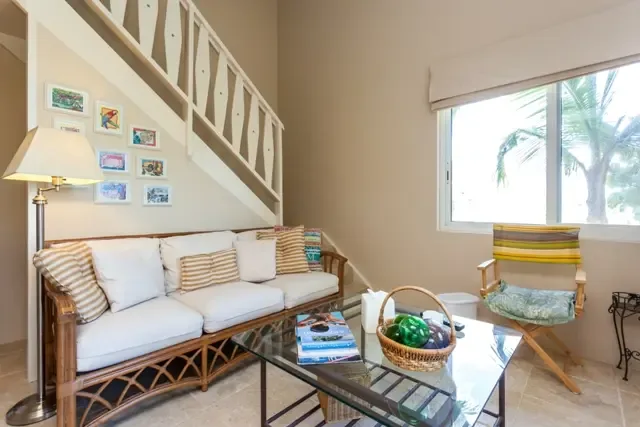 Comfortable island living defines this inviting space, where a wooden-framed sofa and director's chair with striped fabric surround a glass coffee table. A decorative staircase and window views of swaying palm trees create a relaxed Caribbean atmosphere.                                                                                                                                                                                                                                                               +
 