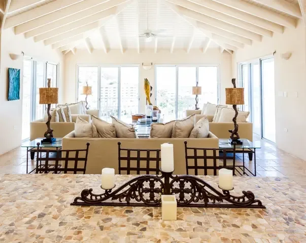 An expansive living room featuring multiple beige sofas, glass side tables, and dark wooden chairs arranged beneath high ceilings. Wicker-shaded floor lamps and an ornate iron candelabra add warmth, while turquoise wall art reflects the Caribbean colors visible through the windows.                                                                                                                                                                                                                                +
 