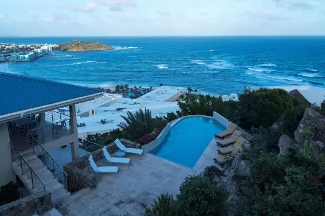 The kidney-shaped pool sparkles with turquoise water beside white and beige lounge chairs on a stone patio. The villa's dark blue roof peeks into frame above lush vegetation, with sweeping views of the ocean and St. Maarten's coastal town below.                                                                                                                                                                                                                                                                       +
 