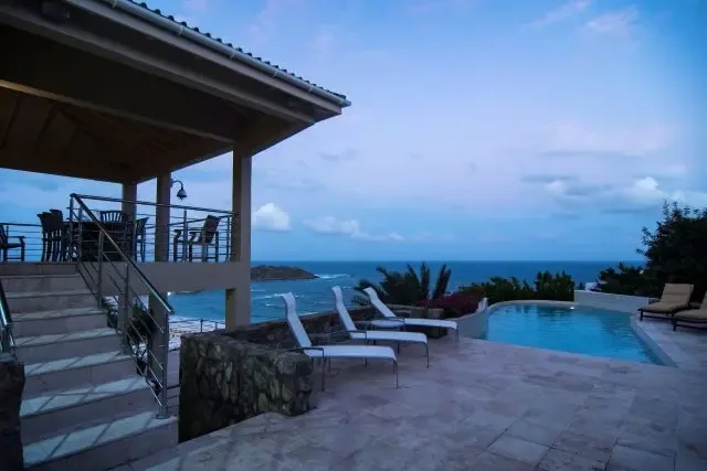 Blue hour beauty at the pool deck, where white lounge chairs line the stone patio beside a covered outdoor dining area. The ocean and distant island create a serene backdrop as the villa's lights begin to twinkle, signaling the transition from day to night in St. Maarten.                                                                                                                                                                                                                                          +
 
