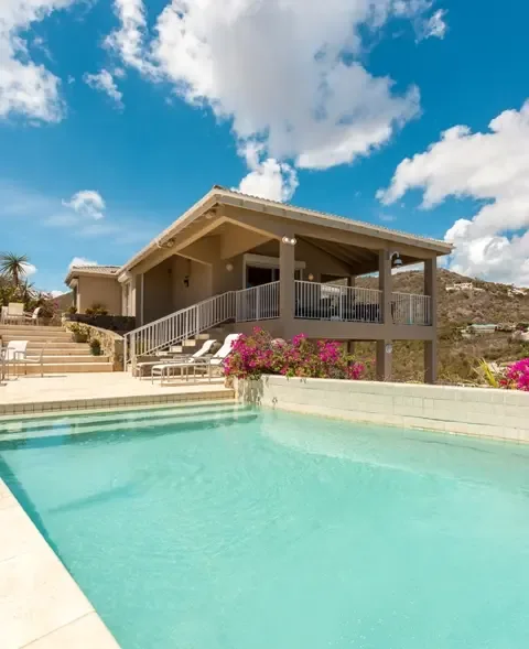 Lounge in paradise beside the rectangular swimming pool, where white lounge chairs and stone staircases with metal railings invite relaxation. Pink bougainvillea cascades beside the villa's covered deck, with St. Maarten's hilly landscape creating a stunning backdrop.                                                                                                                                                                                                                                                  +
 