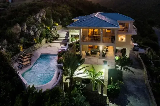 A breathtaking aerial night view of Villa Bougainvillea, where the illuminated infinity pool casts a warm glow across the stone patio. Four lounge chairs await under the Caribbean stars, with lush tropical landscaping and the multi-story villa's warm exterior lighting creating an inviting retreat in Dawn Beach Estates.                                                                                                                                                                                       +
 