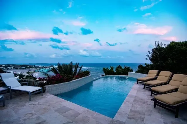 Magic hour at Villa Bougainvillea, where the rectangular pool reflects the twilight sky as purple flowering plants frame the scene. Lounge chairs await in beige and white, with the ocean and coastal town glowing beneath the sunset—a perfect Caribbean evening.                                                                                                                                                                                                                                                           +
 