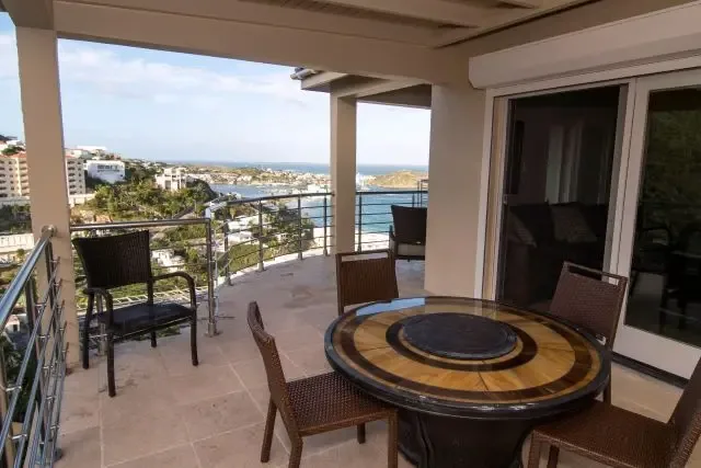 An elegant outdoor dining setting on the covered terrace, with a round decorative table and four wicker chairs positioned to capture panoramic coastal views. Stainless steel railings frame vistas of the ocean, coastal town, and distant hills—a perfect spot for sunset dinners.                                                                                                                                                                                                                                       +
 