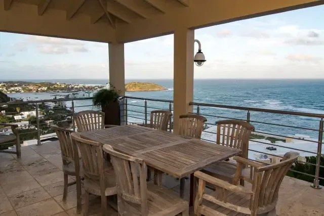 Host unforgettable outdoor feasts at this covered terrace featuring a large wooden dining table with eight matching armchairs. Potted tropical plants and stainless steel railings frame expansive ocean views, with the coastal town and distant island completing the scene.                                                                                                                                                                                                                                             +
 