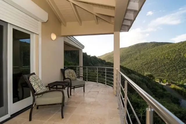 A covered terrace featuring three cushioned armchairs and a side table, perfect for enjoying St. Maarten's mountain and valley views. Stainless steel railings and sliding doors connect this outdoor living space to the villa's interior.                                                                                                                                                                                                                                                                            +
 
