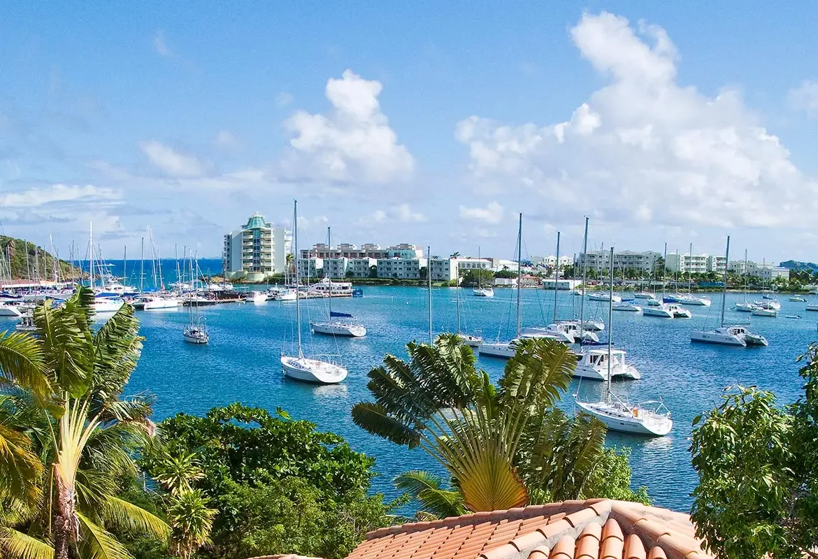 Panoramic harbor view with sailboats and catamarans — the blue bay meets lush tropical greenery and terracotta roofs.