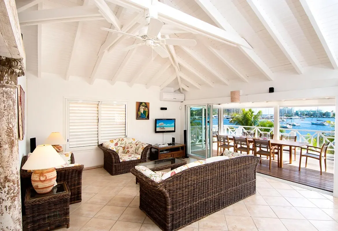Living room opens to a deck with harbor views — vaulted white ceilings and wicker furniture define coastal luxury.
