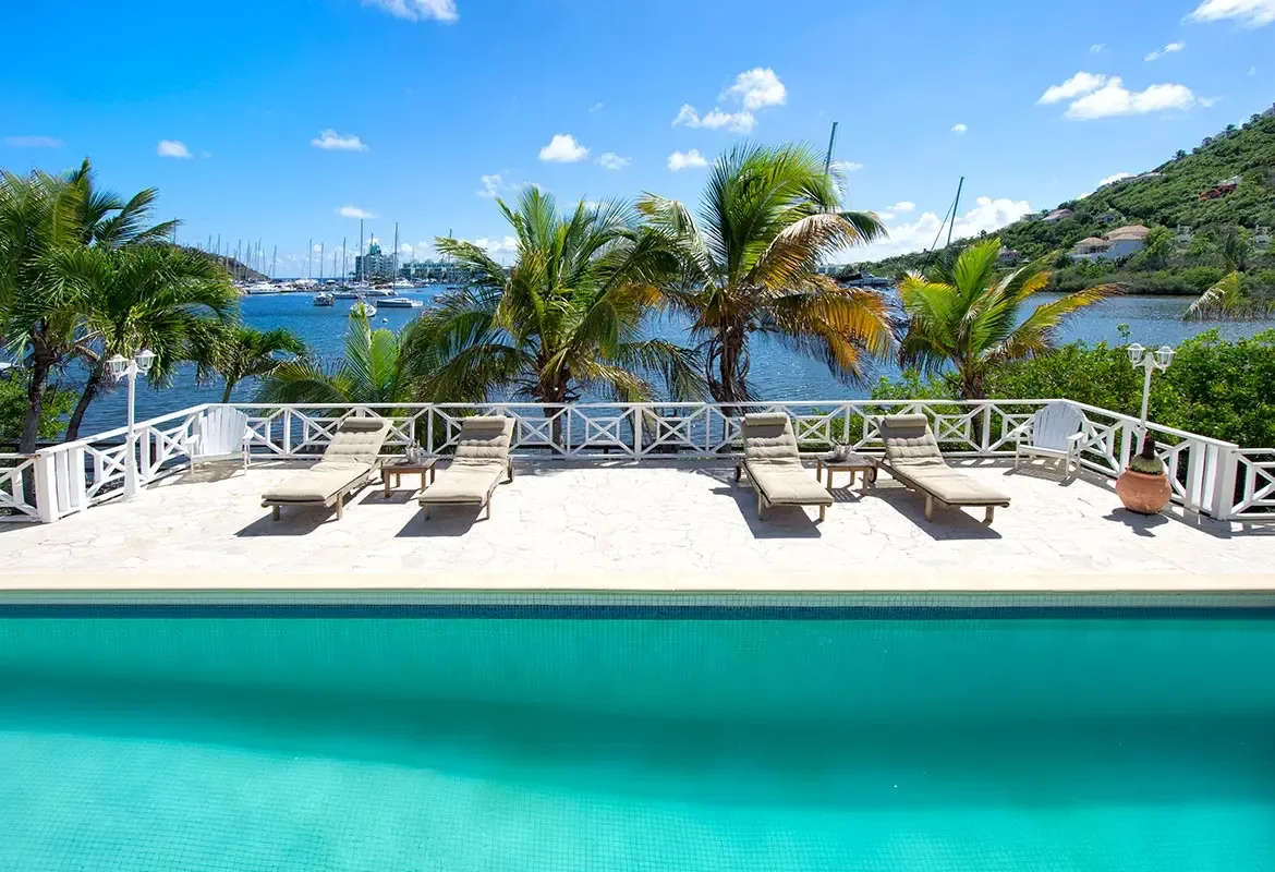 Lounge by the pool with panoramic harbor views — yachts dot the bay while palm trees sway against the hillside.