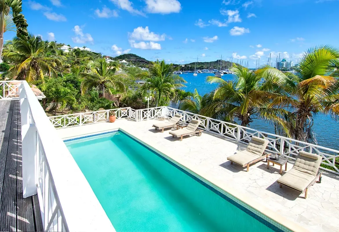 Rectangular pool with lounge chairs and white railing — where turquoise water meets the blue bay and sailboats.