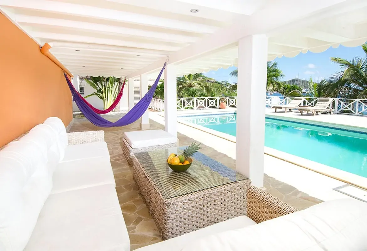 Covered terrace with white sectional sofa and purple hammock — the pool shimmers beyond the tropical foliage.