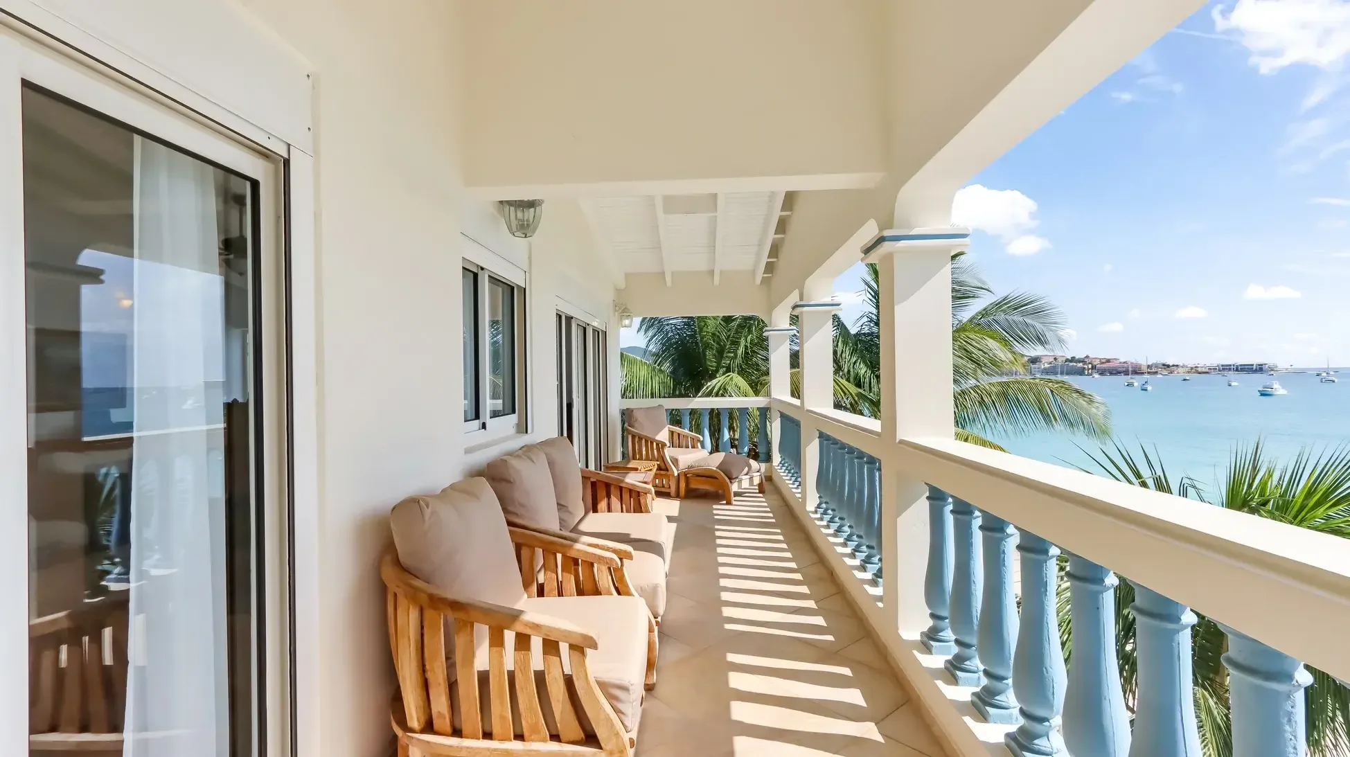 A balcony with wooden armchairs and lounge chair. White columns and blue balustrade frame the tropical bay with turquoise water and boats.