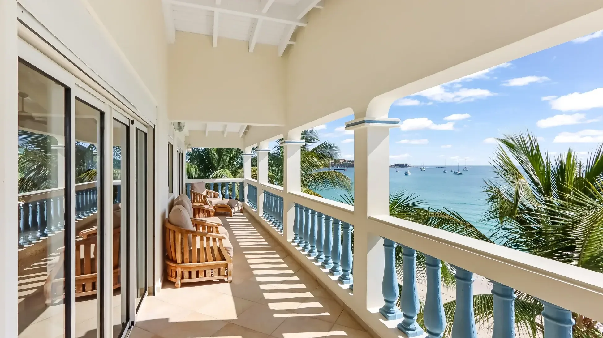 Covered balcony with wooden lounge chairs and cushions. Sliding glass doors open to tropical beach views with palm trees and sailboats.