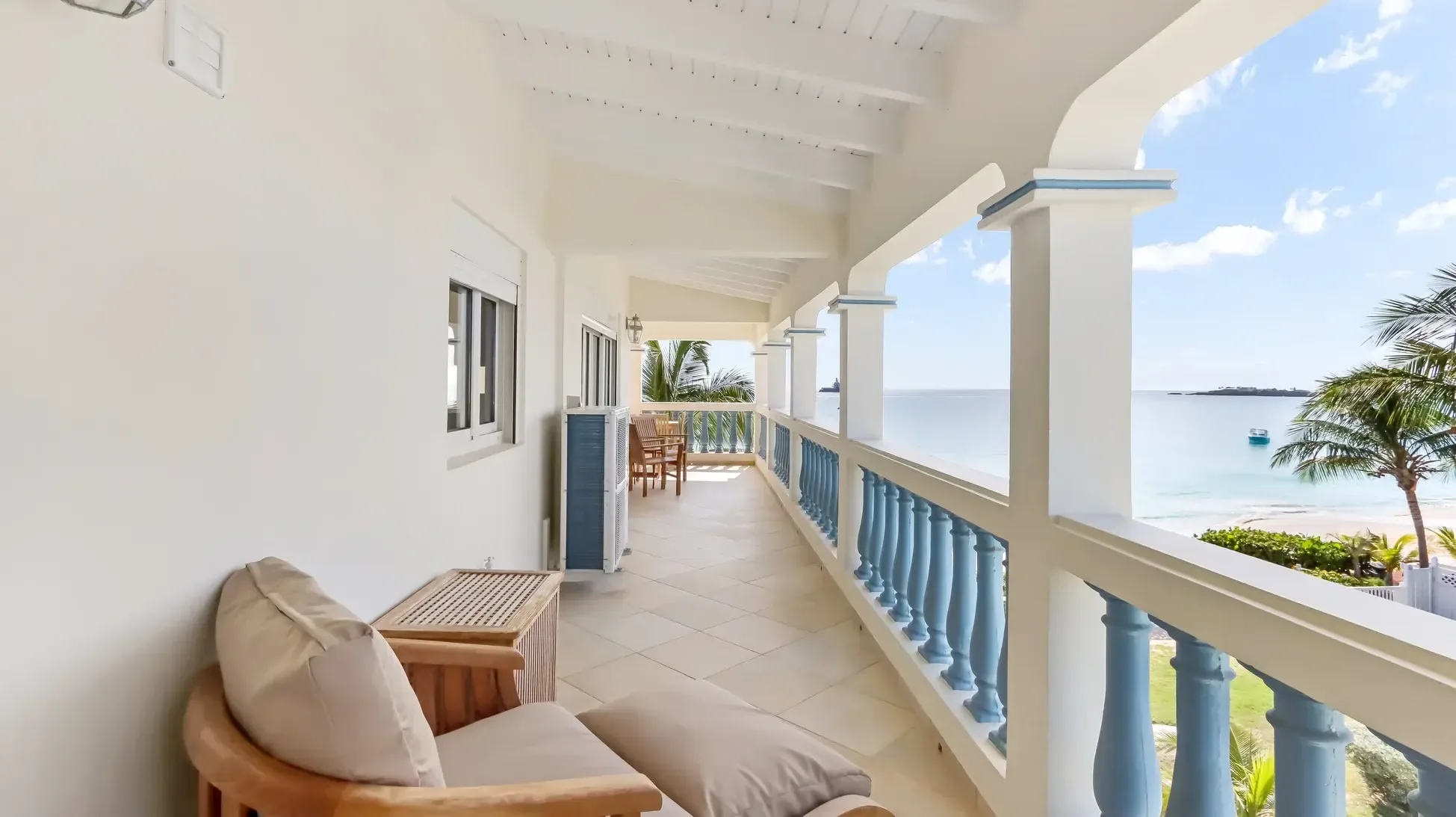 A covered balcony with wooden armchair, ottoman, and dining set. The blue balustrade overlooks the ocean with distant island views.