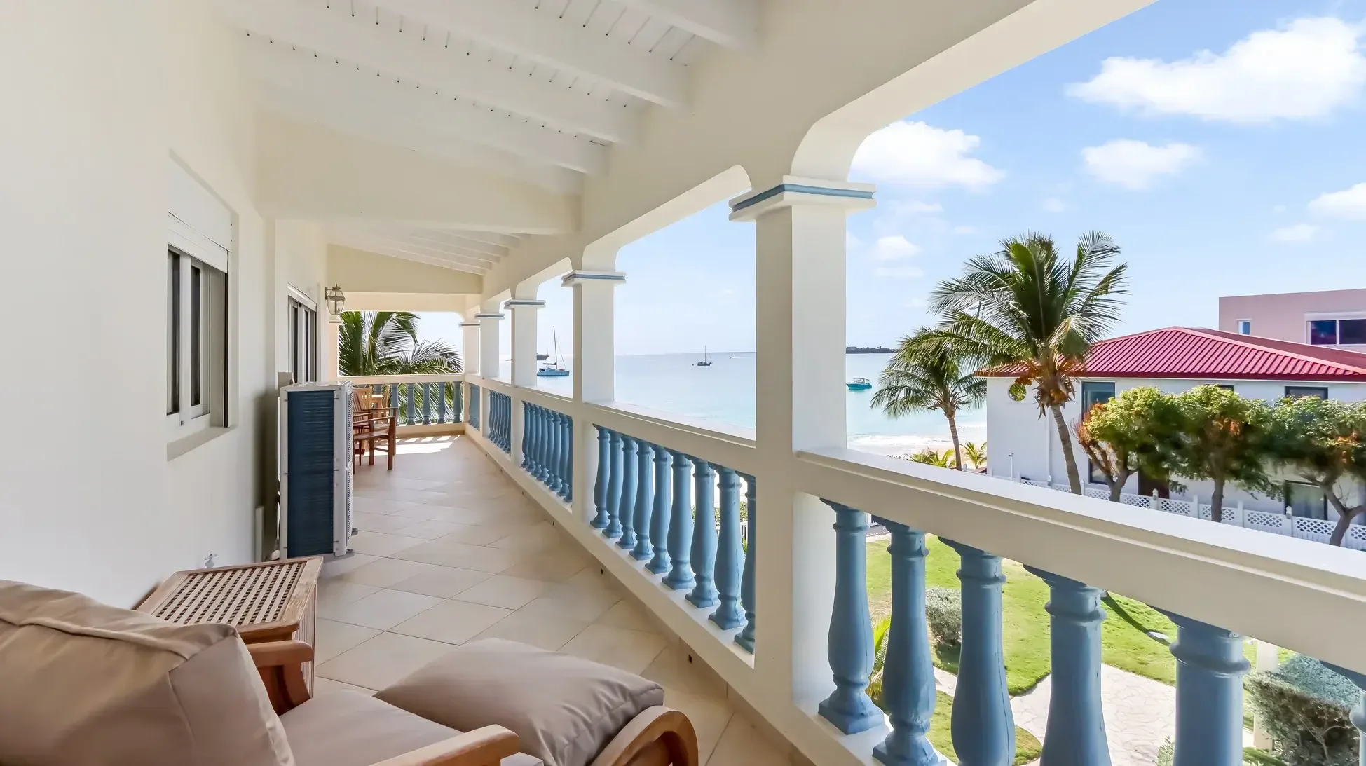 Your covered balcony features cushioned lounge seating and wooden dining set. The blue-and-white balustrade frames turquoise ocean views with beach and palm trees.