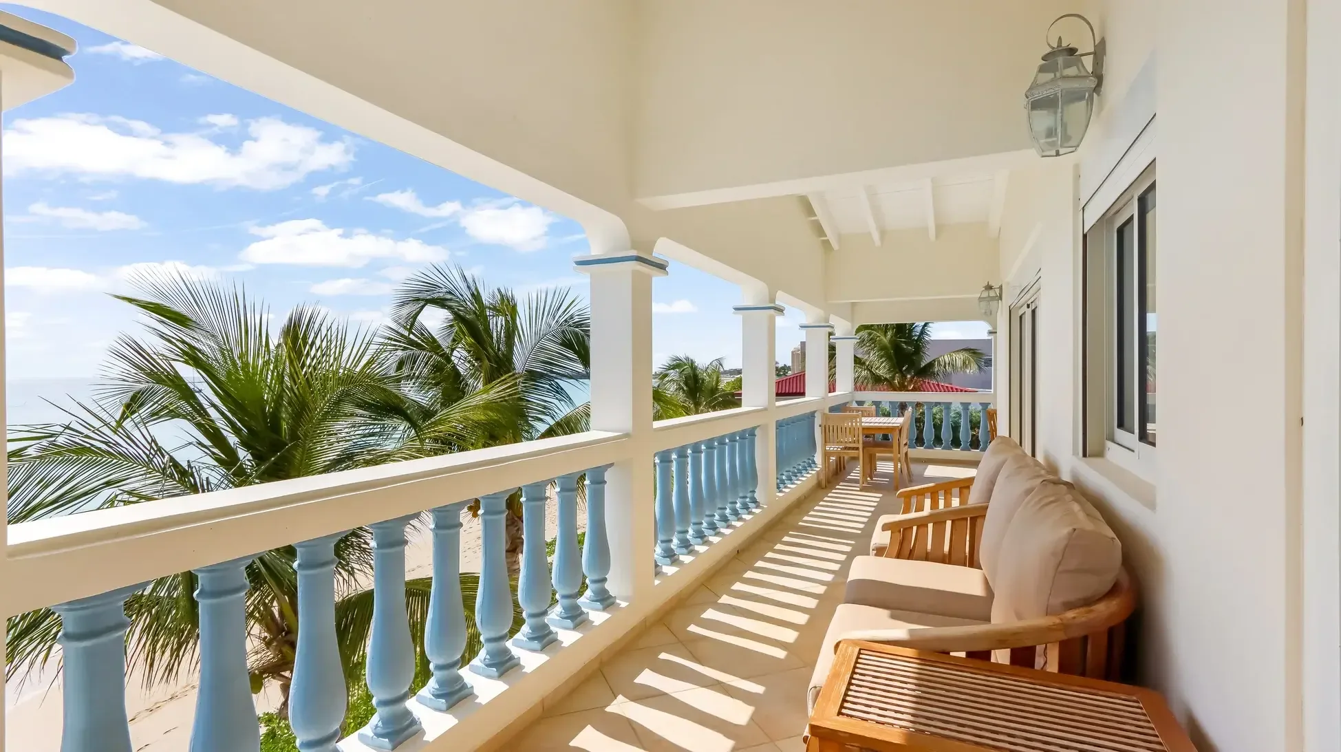 Blue and white balustrade, wooden lounge set with cushions, and dining set frame palm trees and sandy beach ocean views.